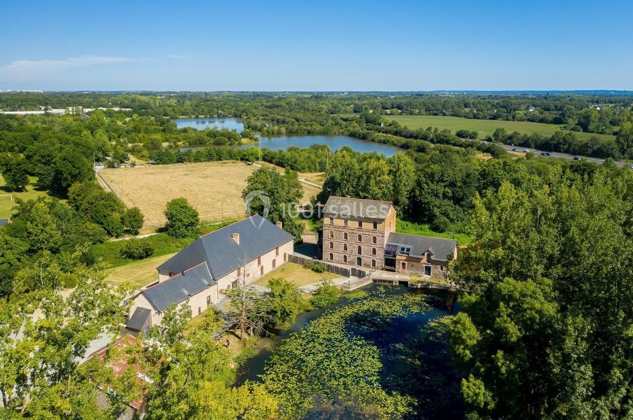 Vue aérienne d'un moulin restauré avec étang, entouré de prairies, de forêts et de plans d'eau sous un ciel dégagé.