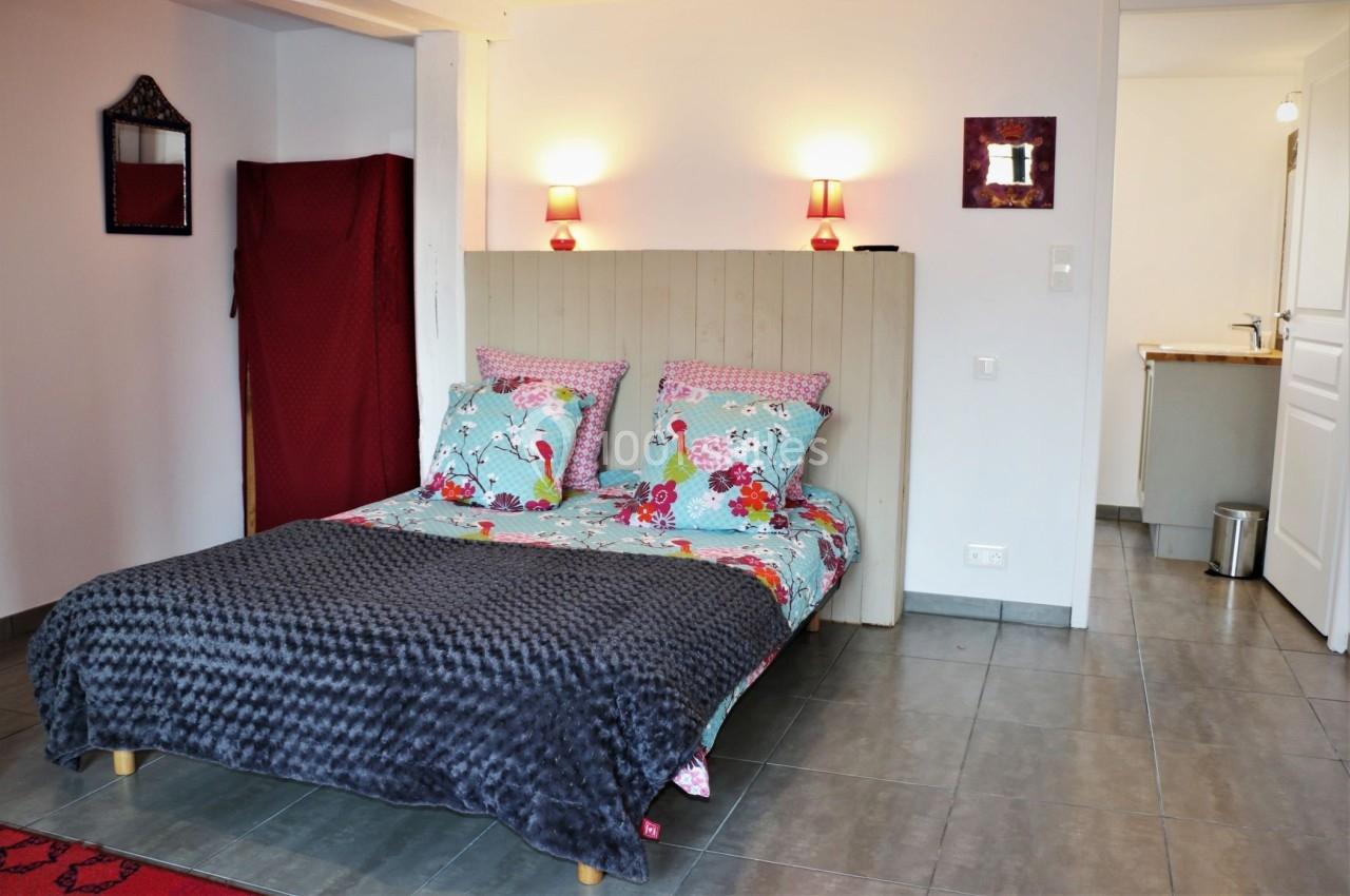 Chambre avec lit double, couvre-lit gris, coussins colorés, murs blancs, éclairage mural et sol en carrelage gris.