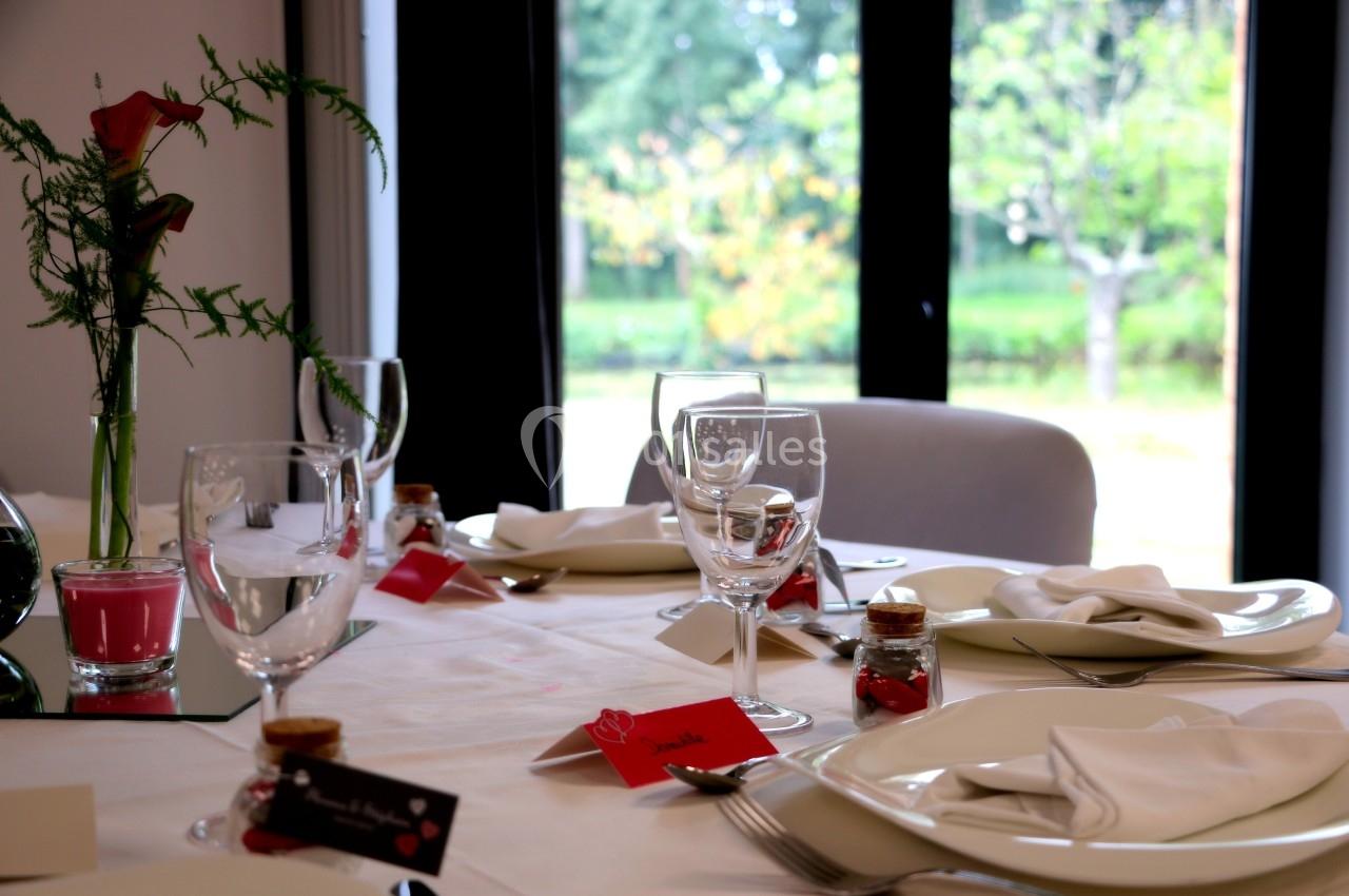 Table dressée avec nappes blanches, verres, serviettes pliées, marque-places rouges et vue sur un jardin verdoyant.