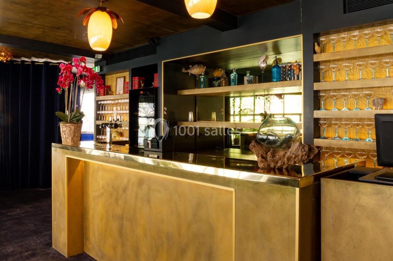 Comptoir doré dans un bar élégant avec étagères de verres, orchidée rouge et éclairage chaleureux.