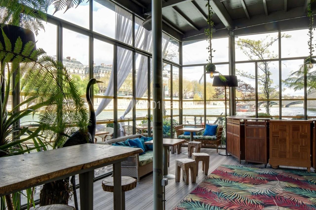 Intérieur lumineux d'une véranda avec mobilier en bois, plantes tropicales et vue sur un paysage urbain.