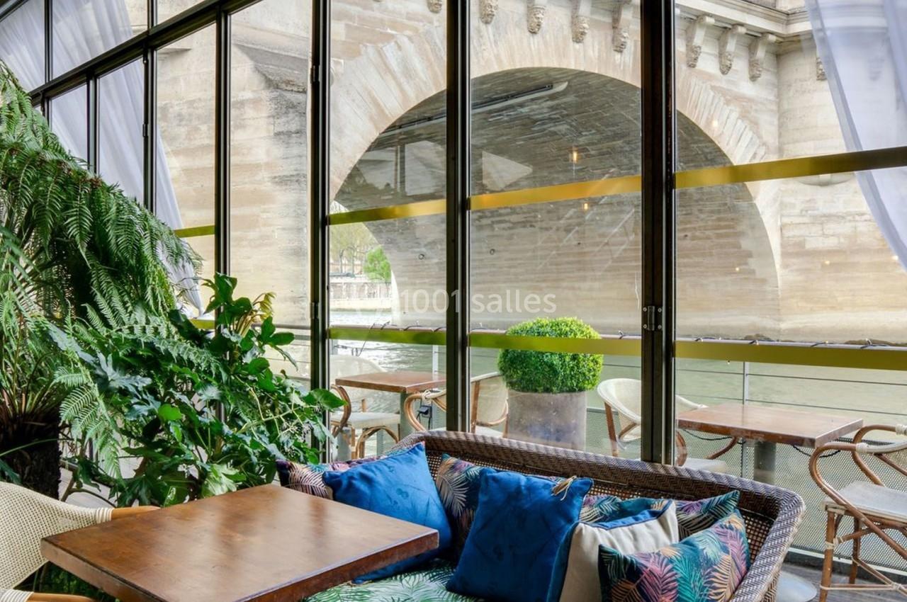 Espace de détente avec fauteuils et plantes, vue sur un pont en pierre à travers de grandes baies vitrées.
