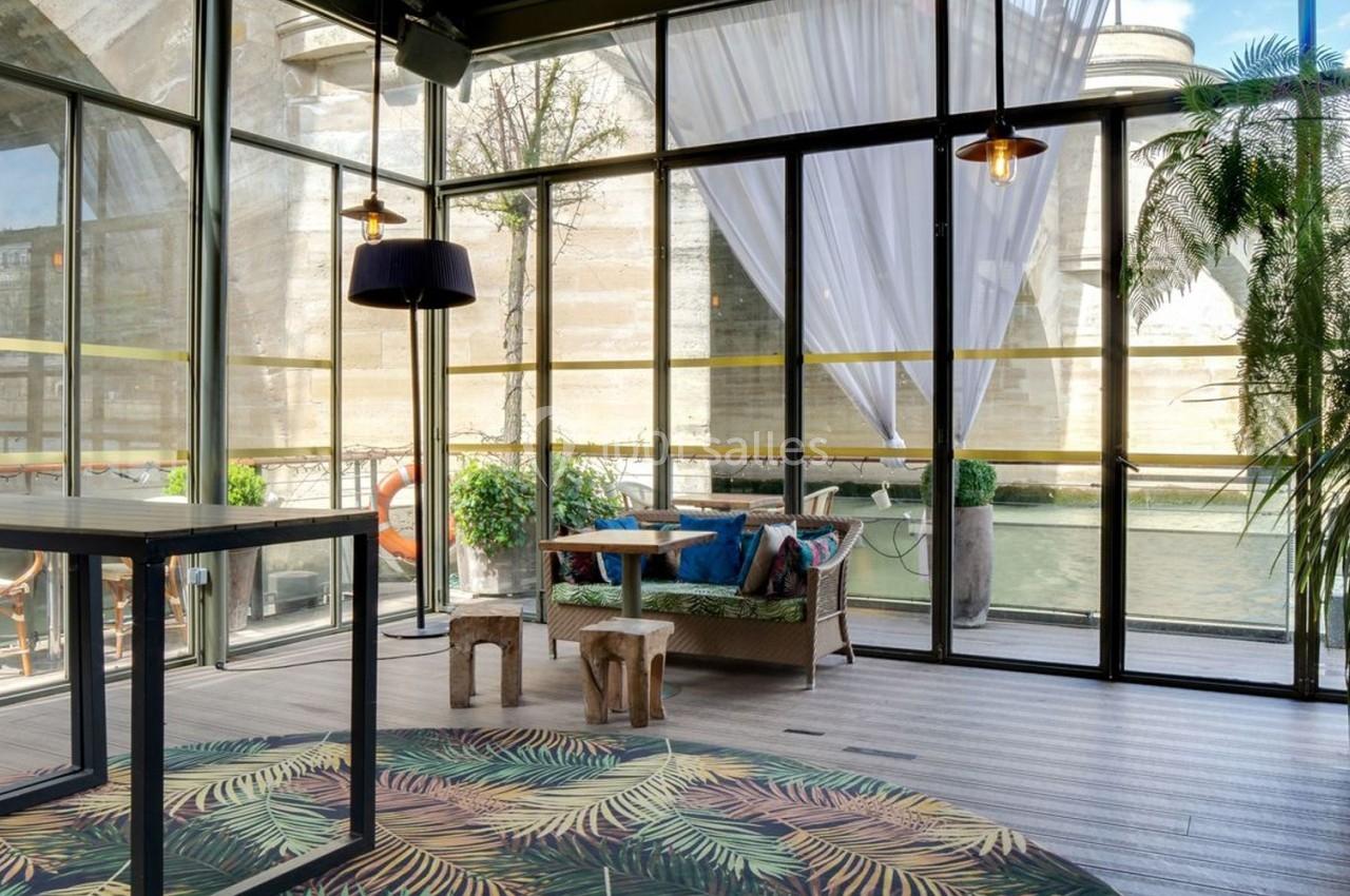 Espace lumineux avec baies vitrées, mobilier en bois, plantes et vue sur un pont en pierre au bord de l'eau.