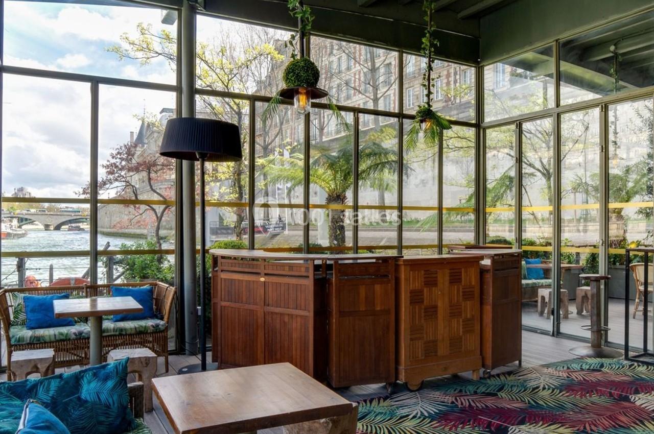 Intérieur d'une véranda lumineuse avec mobilier en bois, plantes suspendues et vue sur un fleuve à travers de grandes baies…