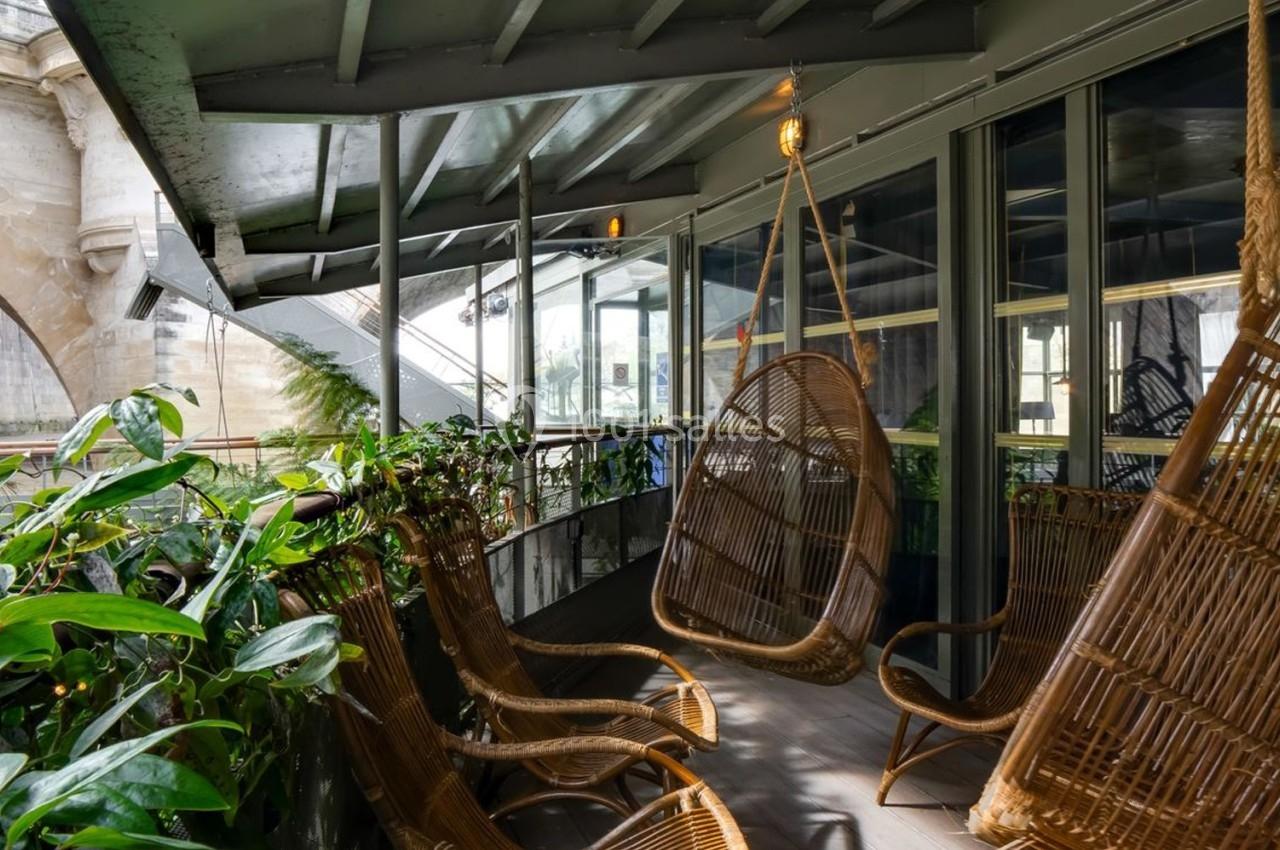 Fauteuils suspendus en rotin sur une terrasse couverte, entourée de plantes, avec vue sur un pont en arrière-plan.