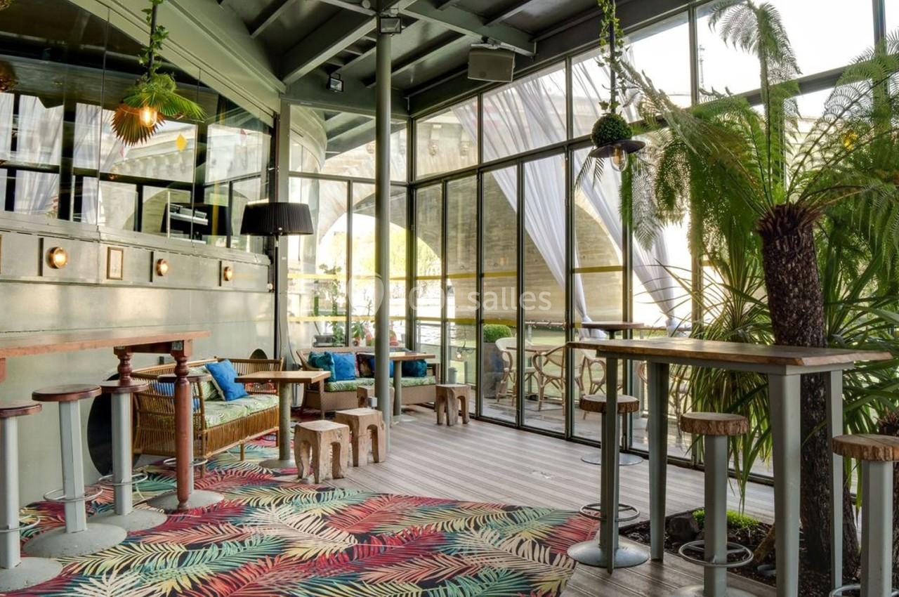 Espace intérieur lumineux avec tables hautes, plantes tropicales et grandes baies vitrées donnant sur l'extérieur.