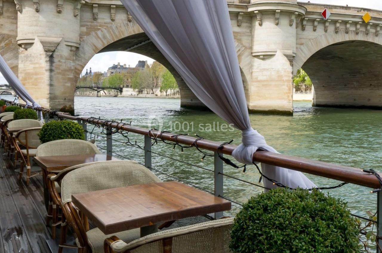 Terrasse avec tables et chaises en bois bordant la Seine, près d'un pont en pierre décoré de drapeaux.