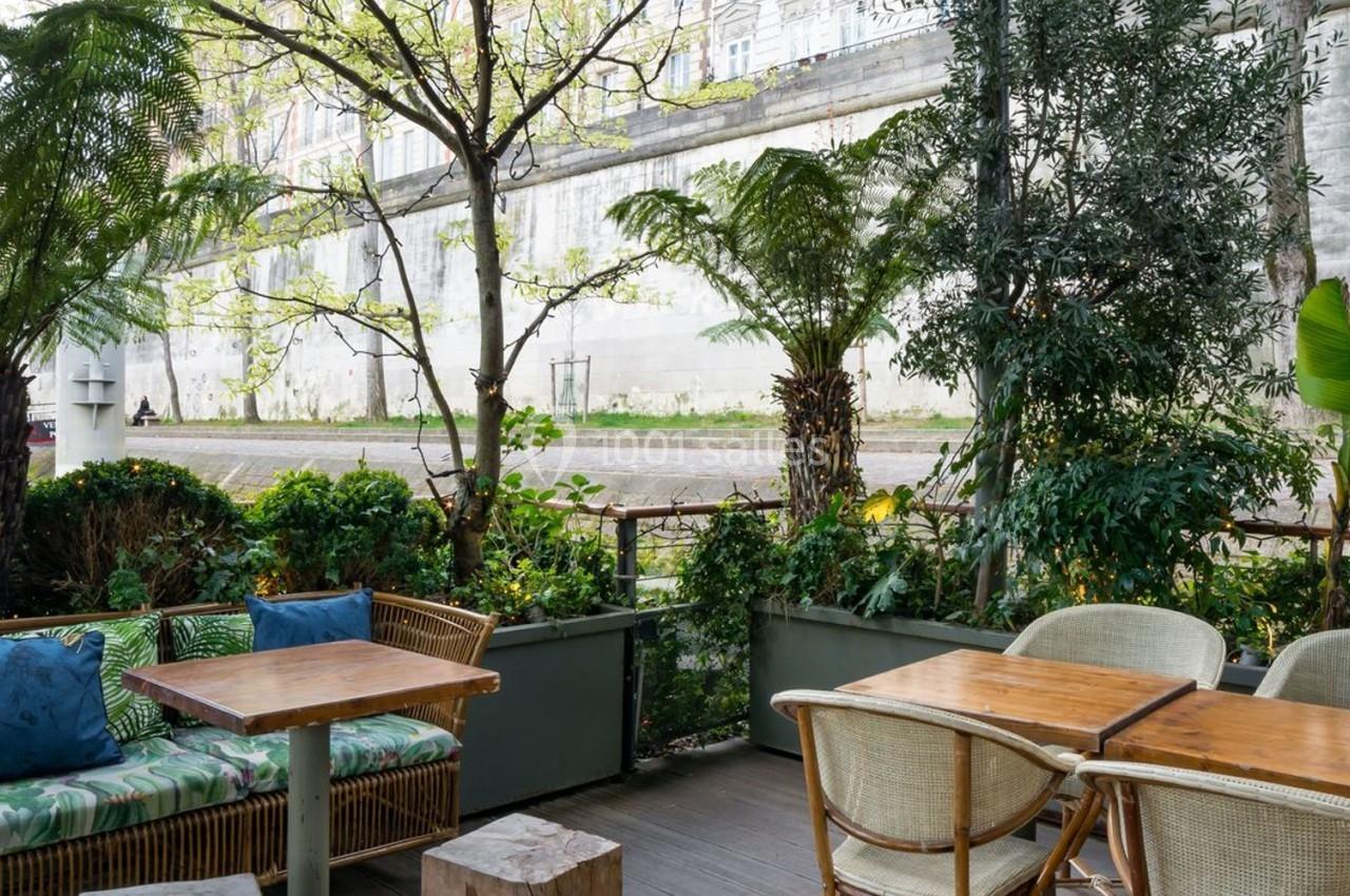 Terrasse extérieure avec tables en bois, chaises en rotin et végétation luxuriante près d'un mur en pierre.