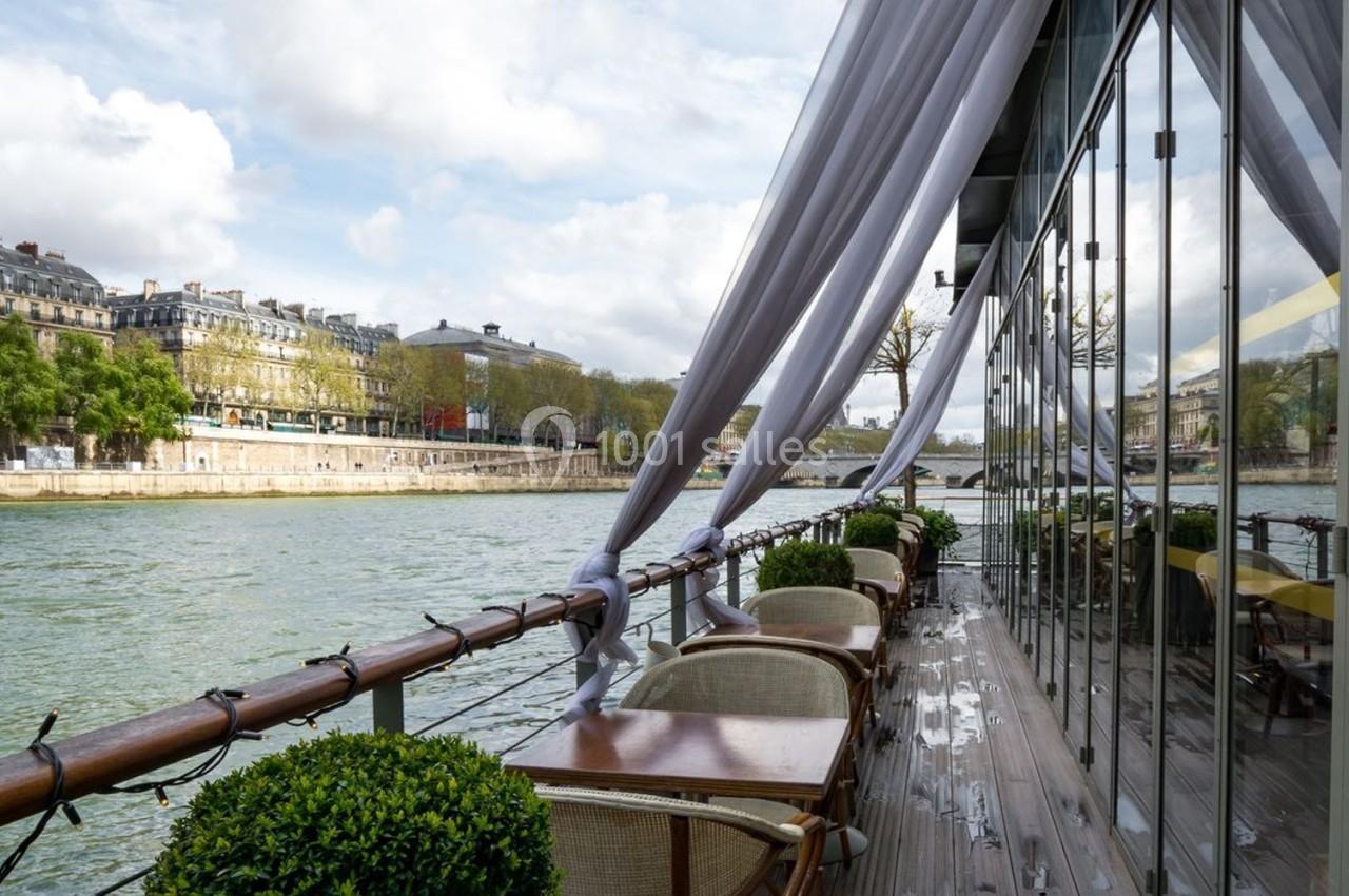Terrasse d'un restaurant flottant avec vue sur la Seine, entourée de végétation et de bâtiments parisiens.