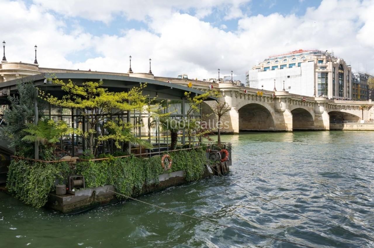 Péniche végétalisée amarrée sur la Seine, près d'un pont en pierre et de bâtiments urbains en arrière-plan.