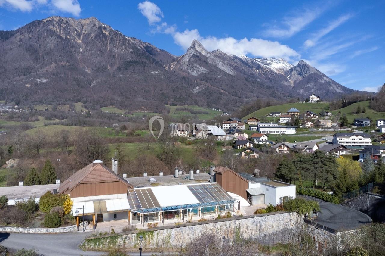 Vue d'un village alpin avec des maisons, une serre et des montagnes en arrière-plan sous un ciel bleu.