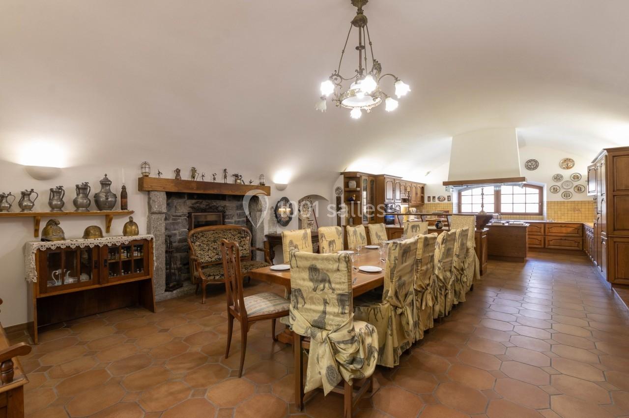 Salle à manger rustique avec table longue, chaises décorées, cheminée en pierre et cuisine ouverte en arrière-plan.