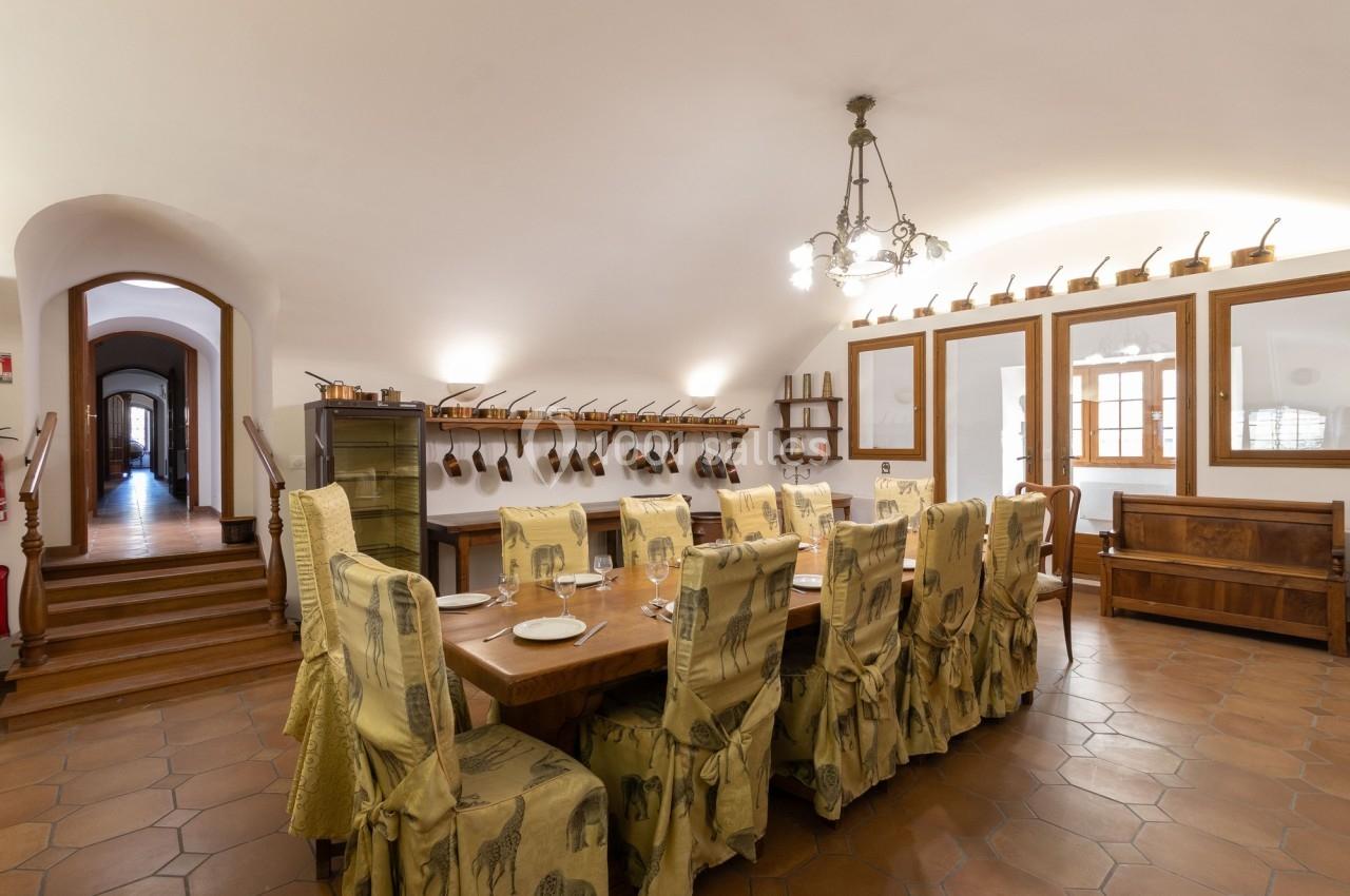 Grande salle à manger avec une longue table en bois, chaises décorées, murs ornés d'ustensiles et éclairage suspendu.