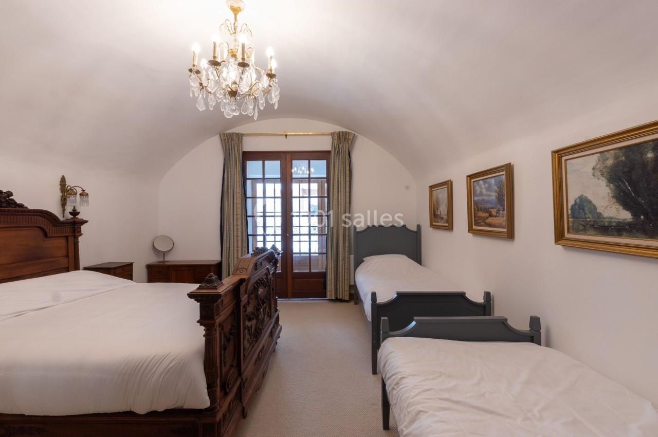 Chambre lumineuse avec un lit double, deux lits simples, meubles en bois, tableaux et lustre, vue sur une porte-fenêtre.