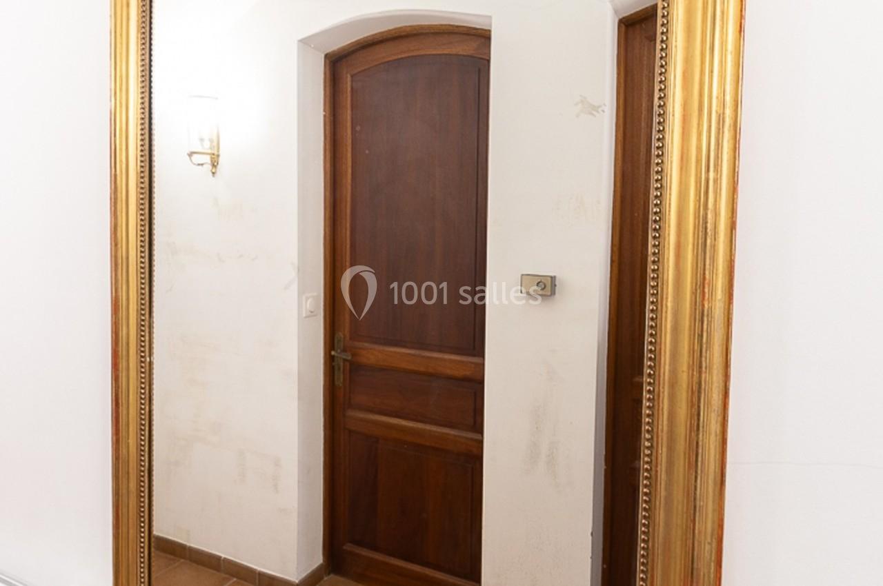 Grand miroir doré reflétant une porte en bois, un mur blanc et un sol en carreaux beige.