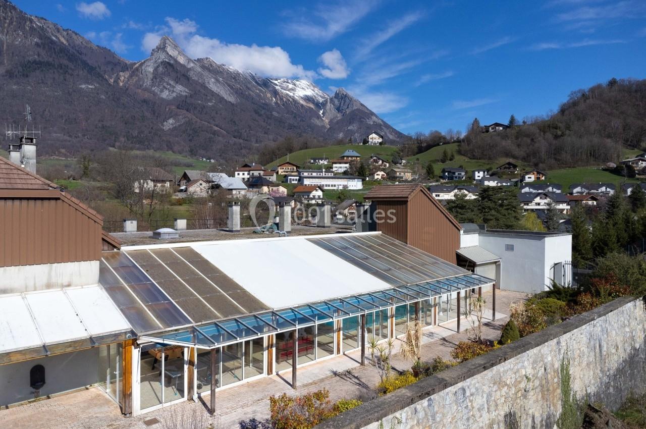 Vue d'une serre vitrée entourée de bâtiments, avec un village et des montagnes en arrière-plan sous un ciel bleu.