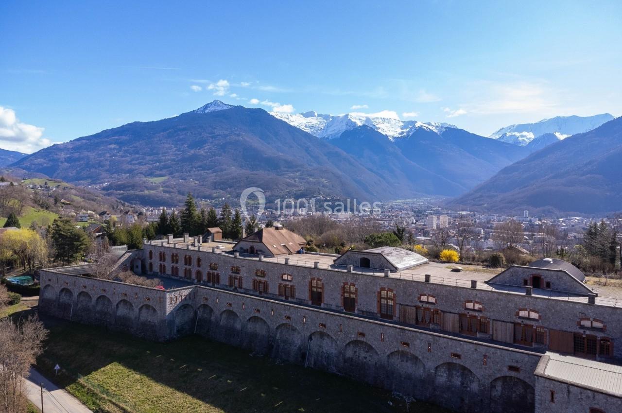 Vue aérienne d'un bâtiment historique en pierre entouré de montagnes enneigées et d'une vallée en arrière-plan.