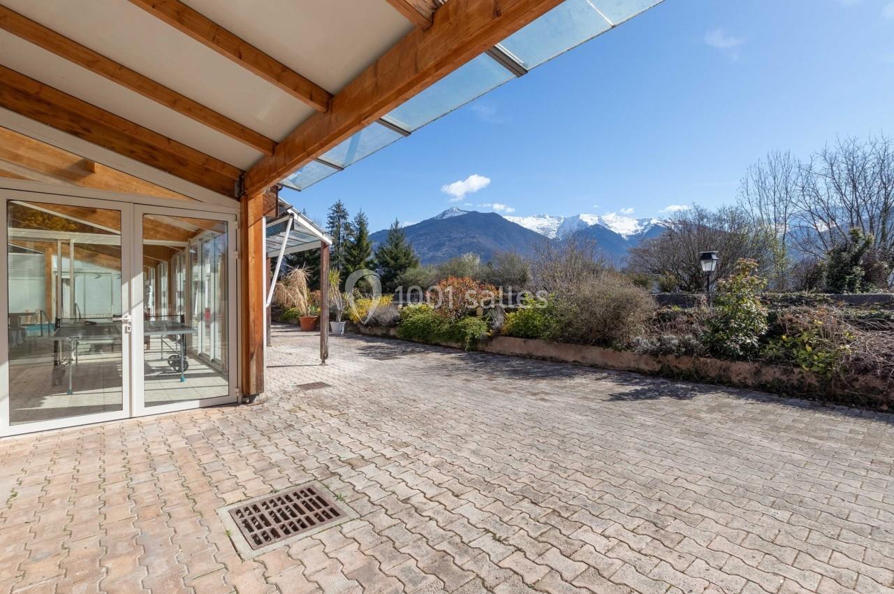 Terrasse pavée avec vue sur des montagnes enneigées, entourée de végétation et d'une véranda en bois et verre.