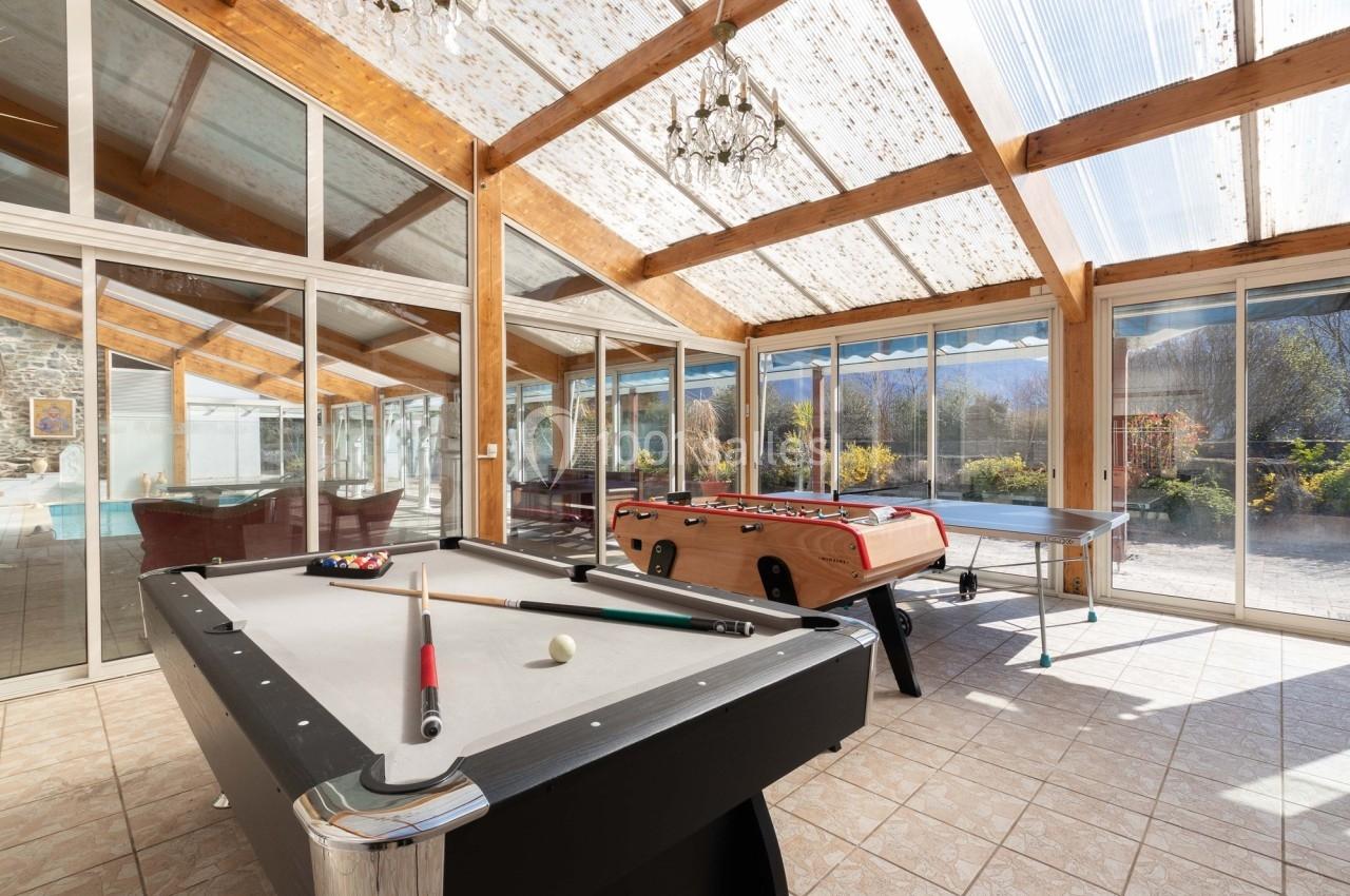 Salle lumineuse avec billard, baby-foot et table de ping-pong, entourée de baies vitrées donnant sur un jardin.
