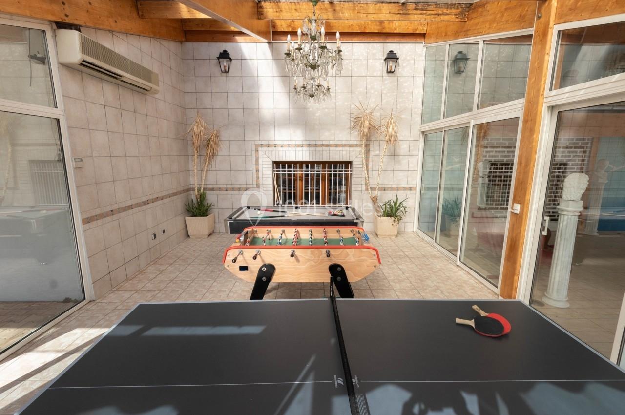 Salle lumineuse avec table de ping-pong, baby-foot et billard, entourée de murs carrelés et de plantes décoratives.