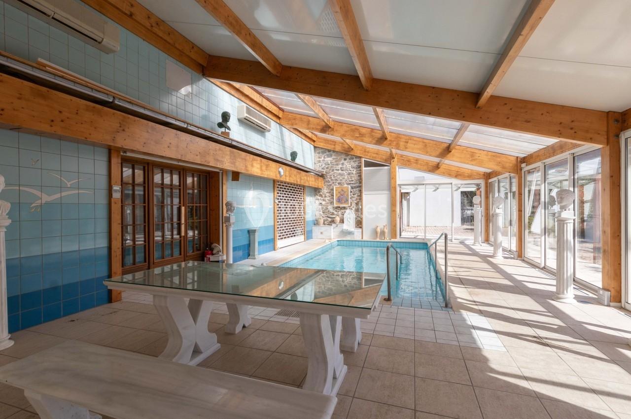 Piscine intérieure lumineuse avec toit en verre, murs en carrelage bleu, table en verre et bancs blancs.