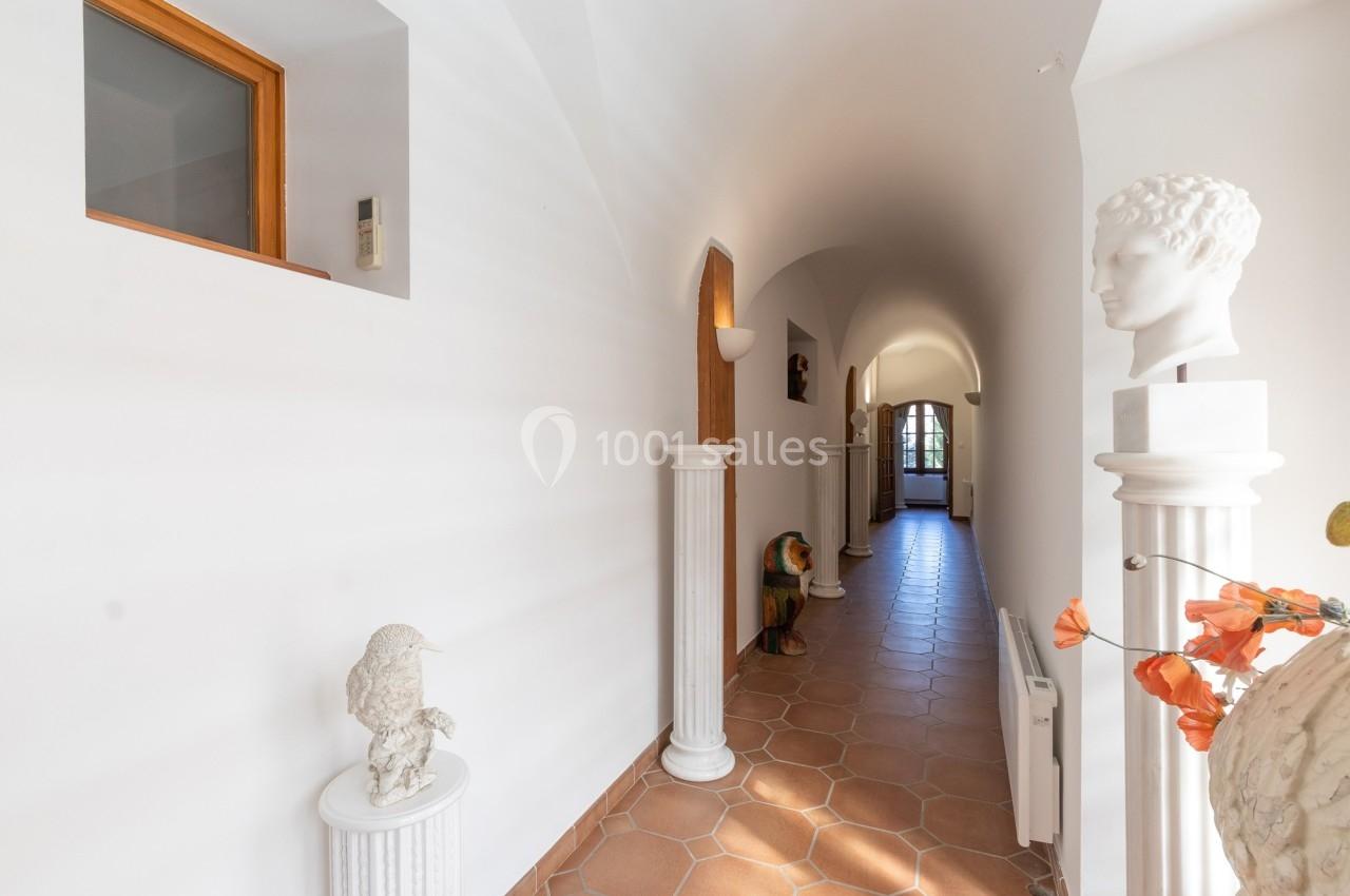 Couloir lumineux avec sol en terre cuite, colonnes décoratives, sculptures et porte vitrée au fond.