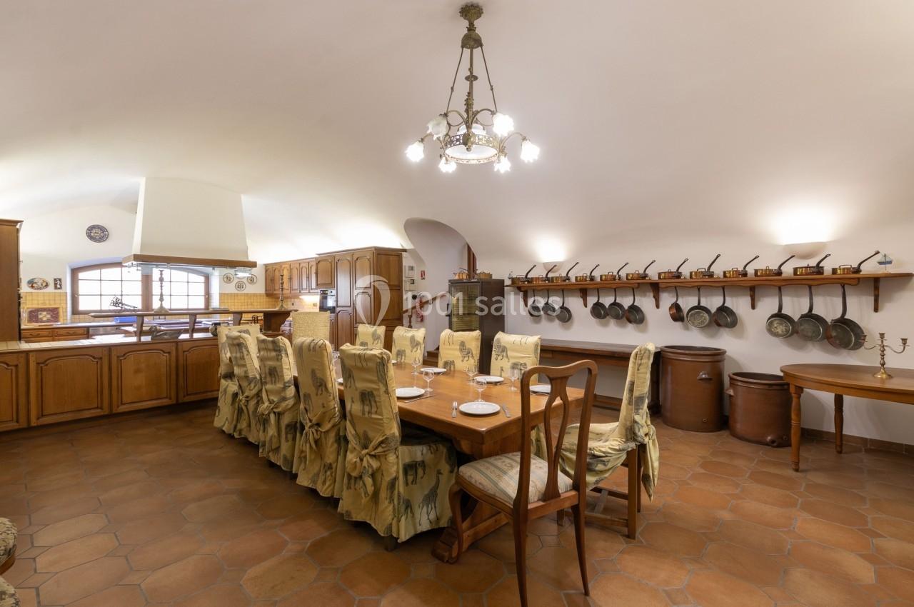 Grande cuisine rustique avec table à manger, chaises décorées, étagère murale avec poêles et carrelage au sol.