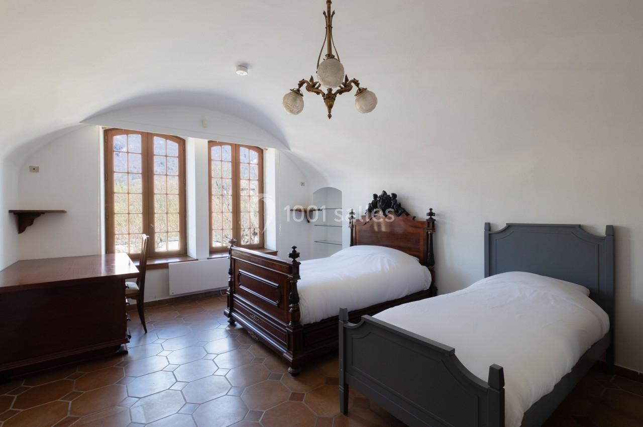 Chambre lumineuse avec deux lits simples, un bureau en bois, un lustre ancien et des fenêtres donnant sur l'extérieur.