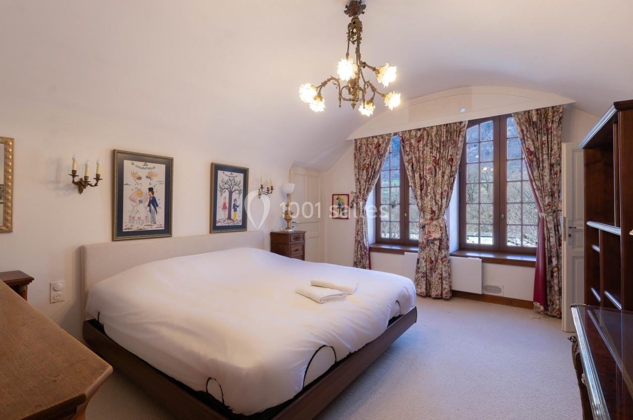 Chambre lumineuse avec un grand lit, des rideaux fleuris, des tableaux encadrés et un lustre ancien.