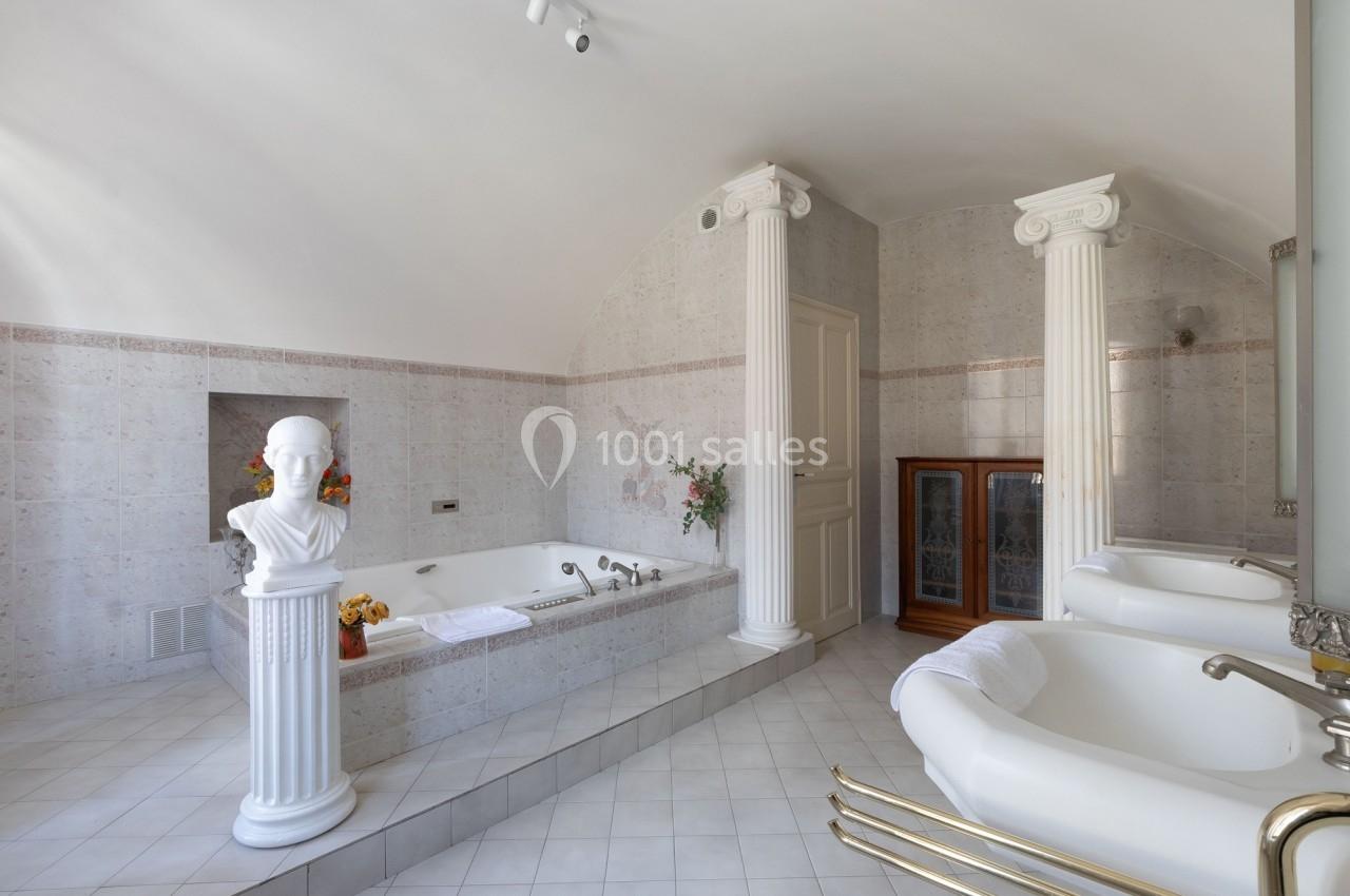 Salle de bain lumineuse avec baignoire encastrée, colonnes décoratives, statue sur socle et lavabos doubles.