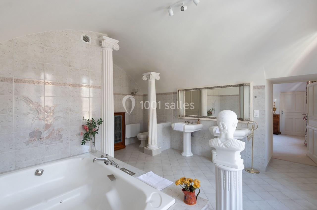 Salle de bain lumineuse avec baignoire, colonnes décoratives, miroir mural et carrelage clair.