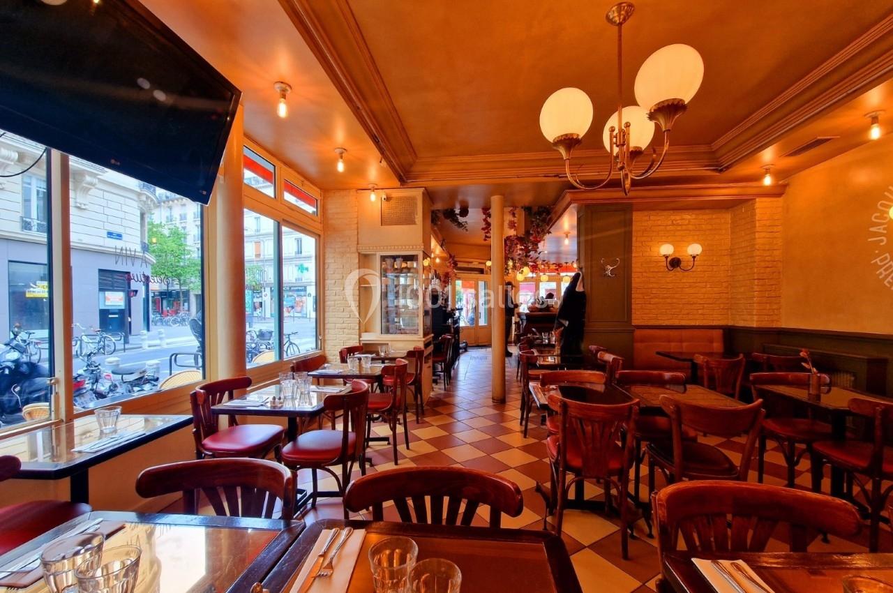 Salle de restaurant avec tables en bois, chaises rouges et grandes fenêtres donnant sur une rue urbaine.