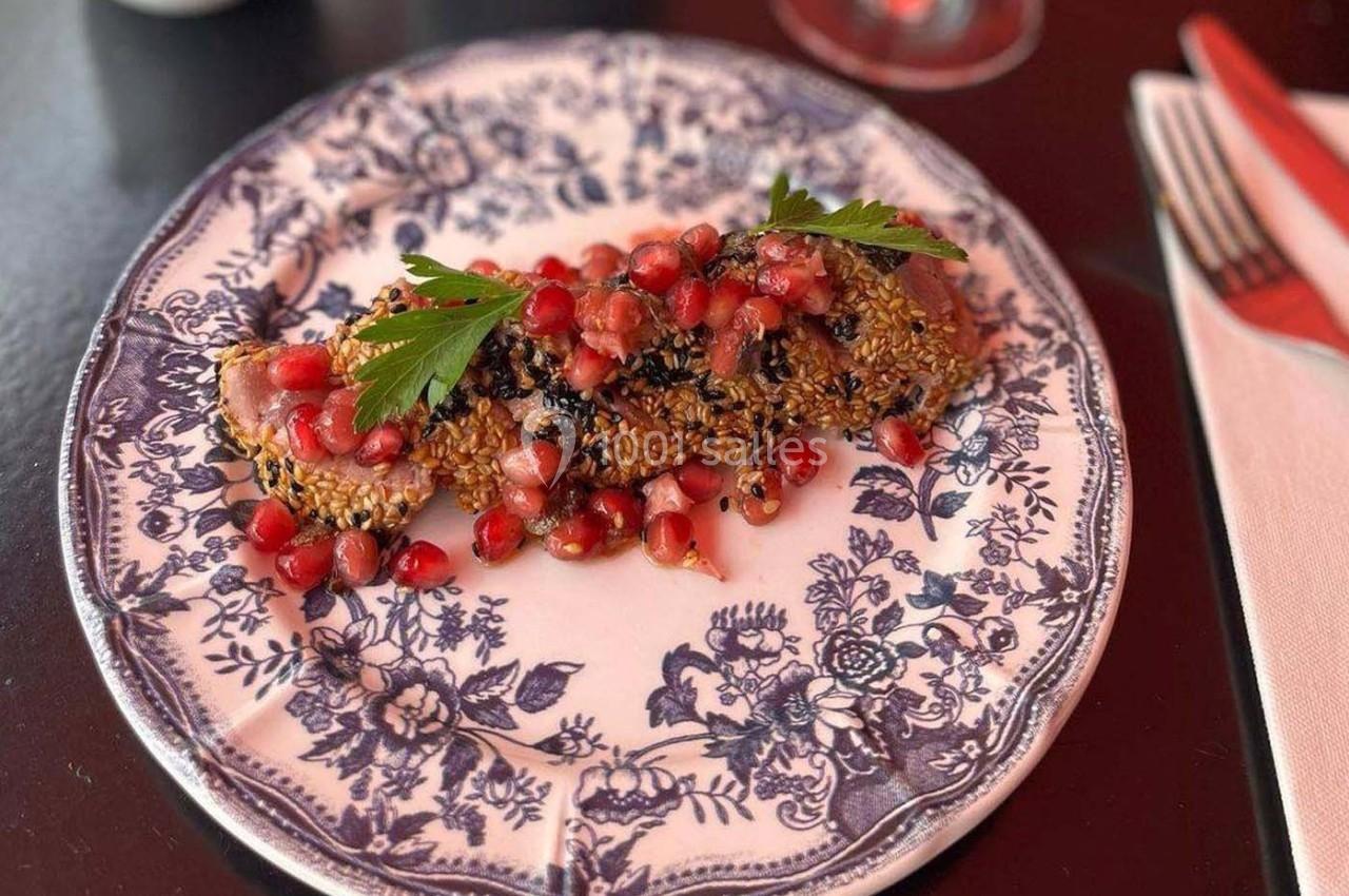 Assiette en porcelaine décorée contenant un plat de saumon en croûte de sésame, garni de grenade et herbes fraîches.