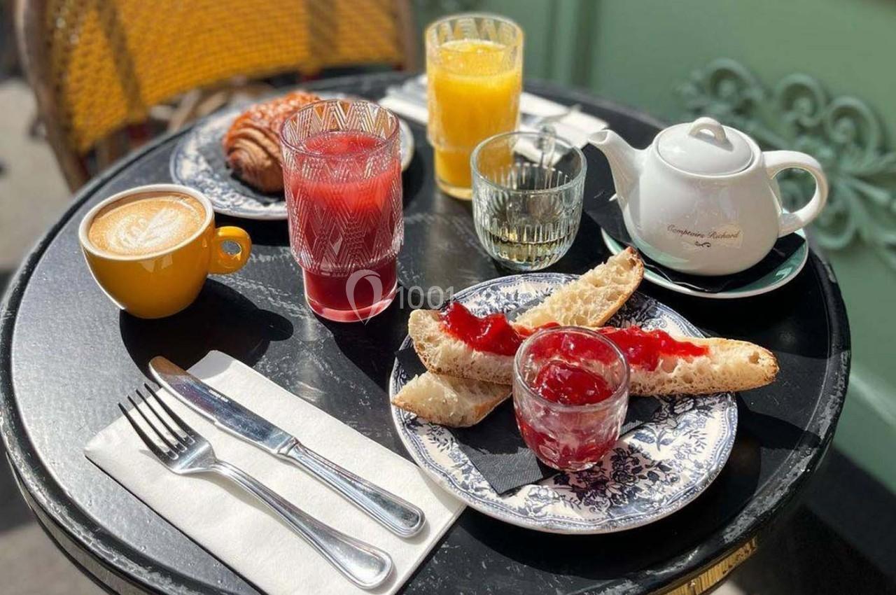 Petit-déjeuner en terrasse avec tartines à la confiture, croissant, boissons variées et une théière blanche.
