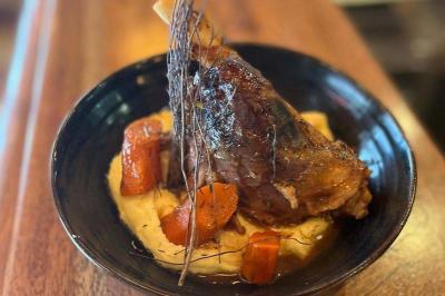 Jarret d'agneau braisé servi sur une purée crémeuse avec des morceaux de carottes, présenté dans une assiette noire.