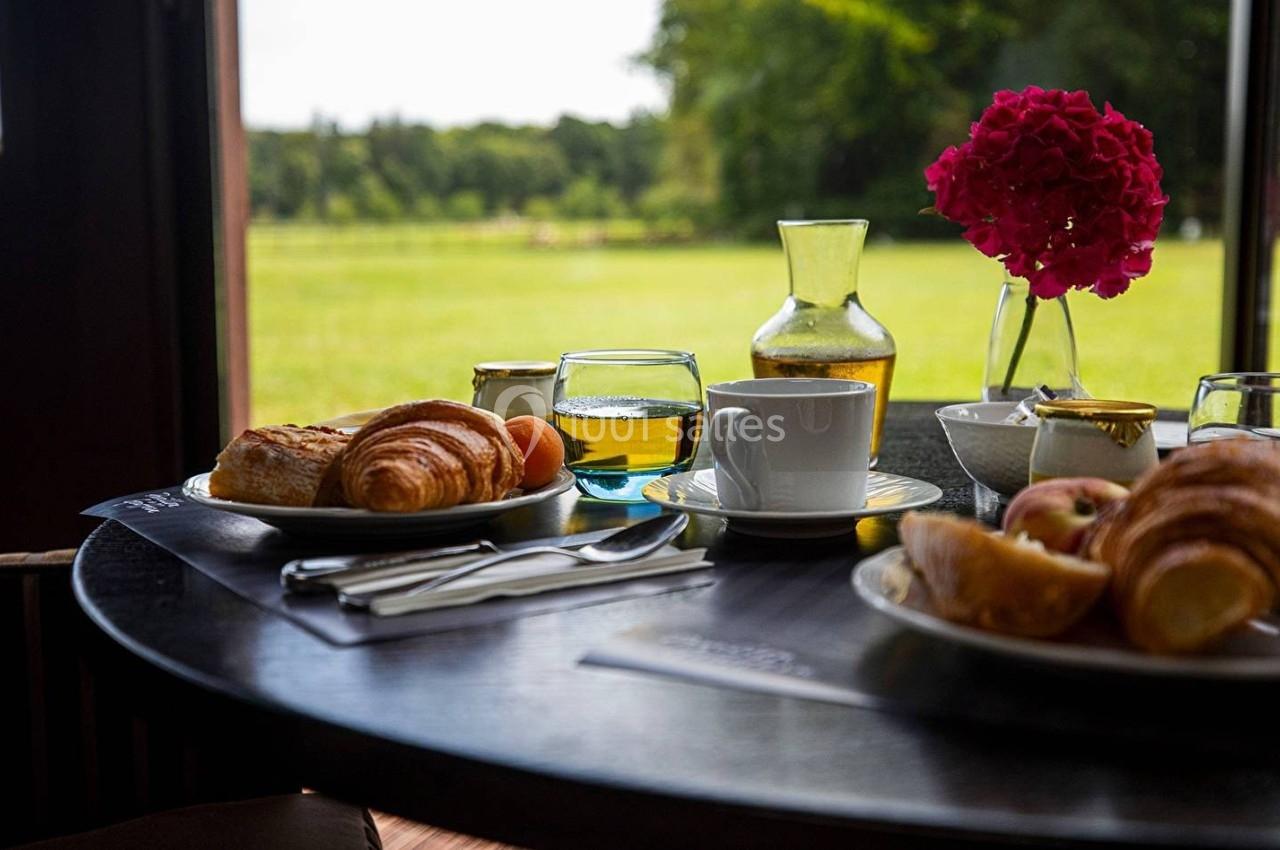 Petit-déjeuner avec croissants, jus d'orange et café sur une table ronde, vue sur un jardin verdoyant en arrière-plan.