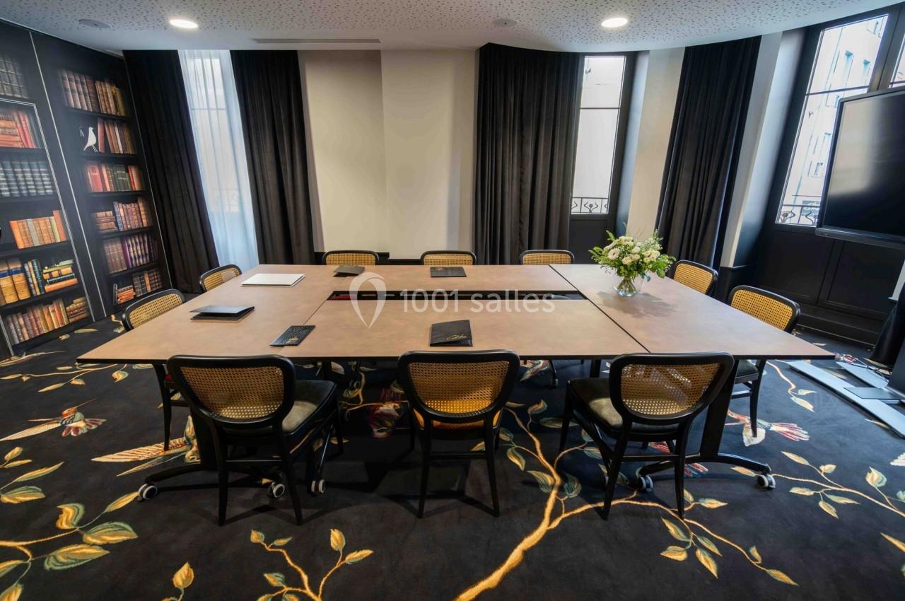Salle de réunion lumineuse avec grande table rectangulaire, chaises en rotin, étagères de livres et décoration florale.