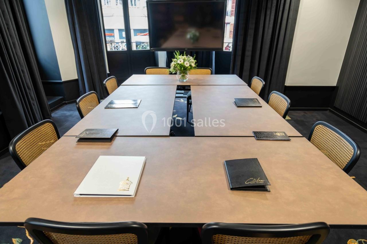 Salle de réunion avec une table en U, des chaises en rotin, des cahiers disposés et un écran en arrière-plan.