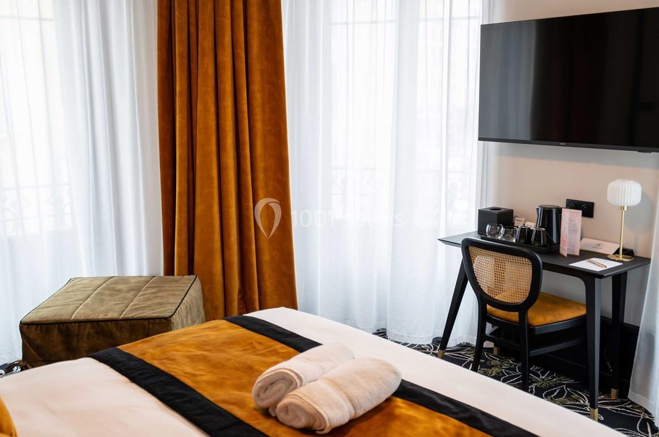 Chambre lumineuse avec lit, bureau équipé, fauteuil, pouf et télévision, décorée de rideaux et textiles couleur ocre.