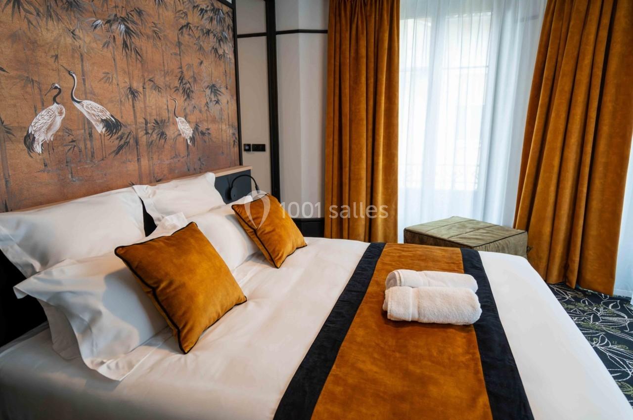 Lit double avec draps blancs, coussins et jeté de lit orange, devant une fenêtre avec rideaux assortis et un décor mural.