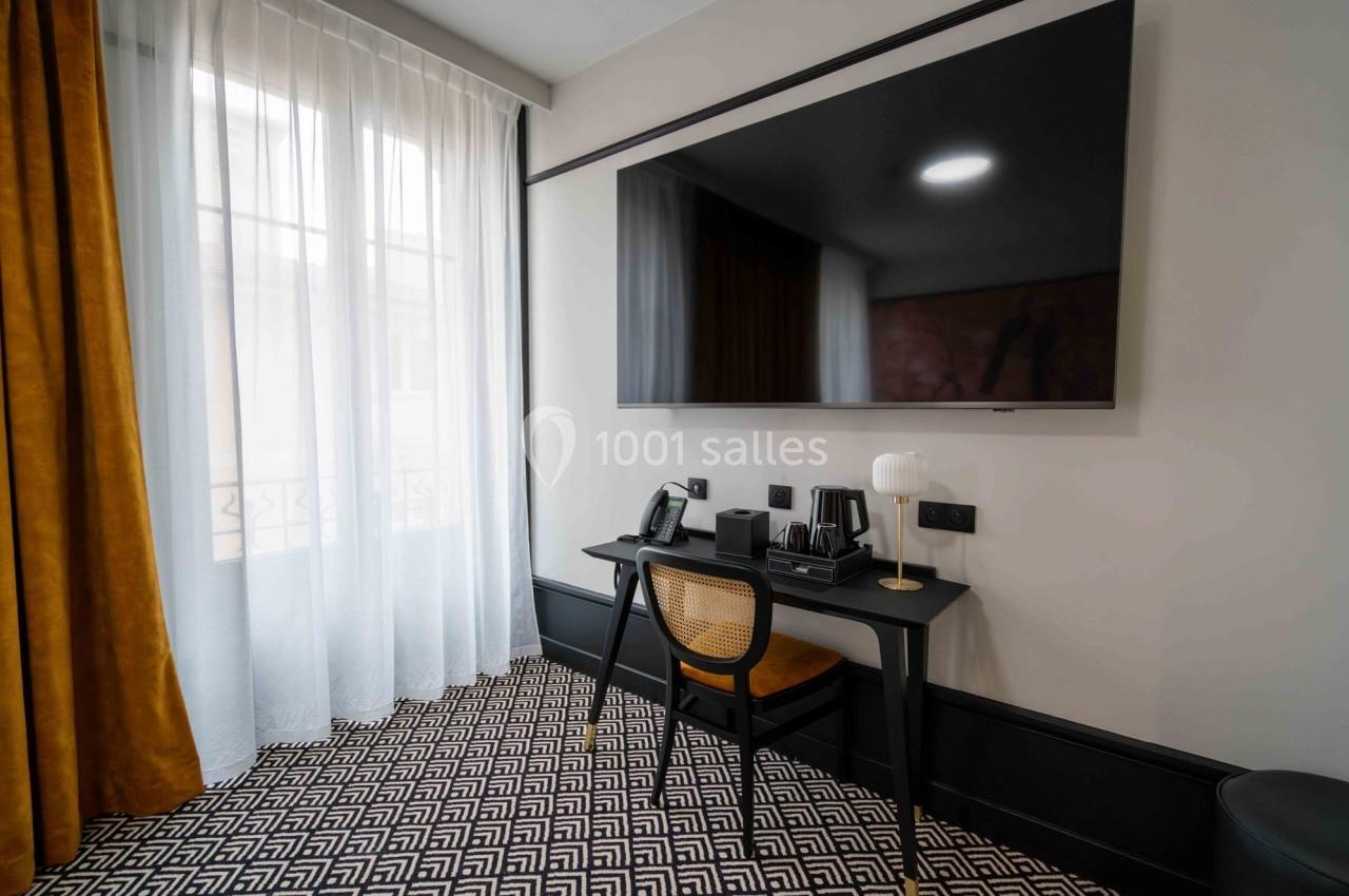 Bureau avec chaise, lampe, téléphone et plateau de courtoisie, face à un grand téléviseur mural dans une pièce lumineuse.