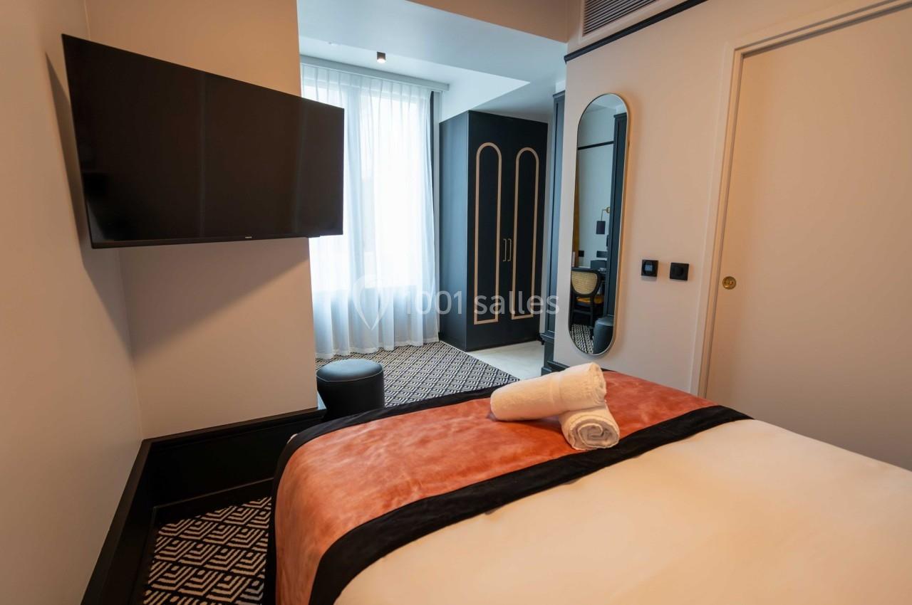 Chambre d'hôtel moderne avec lit double, télévision murale, miroir, fenêtre lumineuse et serviette pliée sur le lit.