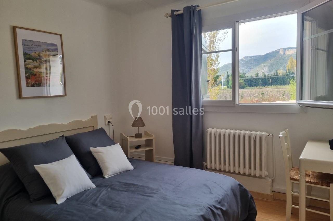 Chambre lumineuse avec lit double, bureau, fenêtre ouverte donnant sur un paysage de montagne et rideaux gris.