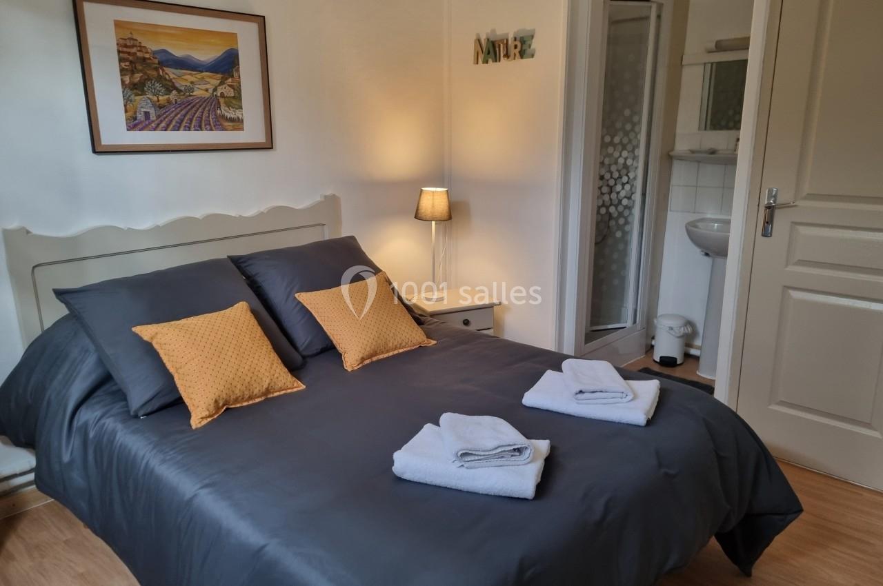 Chambre avec lit double, linge de lit gris, coussins jaunes, lampe de chevet et salle de bain attenante.