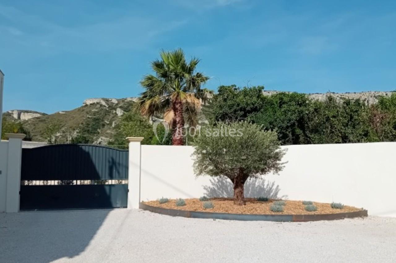 Cour avec portail fermé, mur blanc, palmier, olivier et massif de plantes, située devant un paysage de collines.