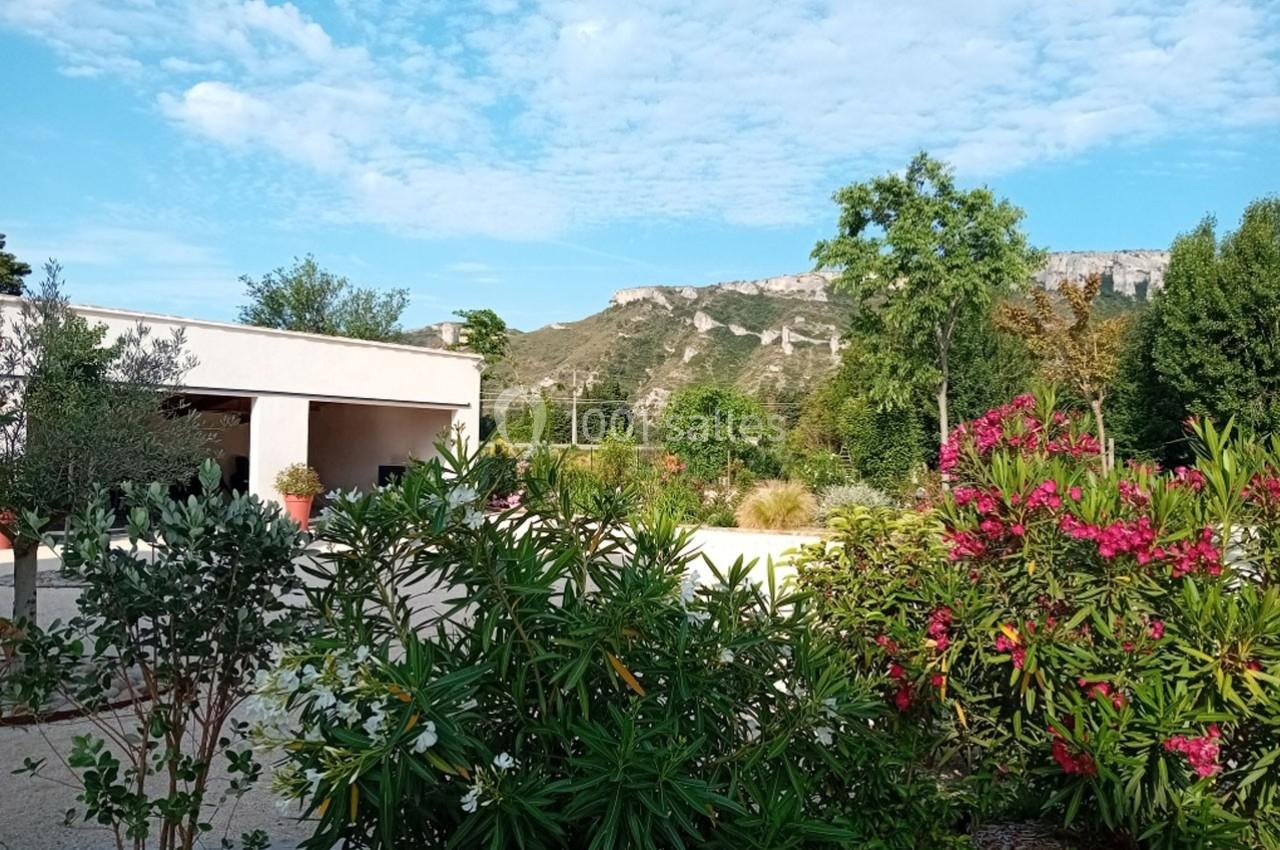 Jardin avec des lauriers roses, arbres et végétation, devant un bâtiment blanc et des collines en arrière-plan.