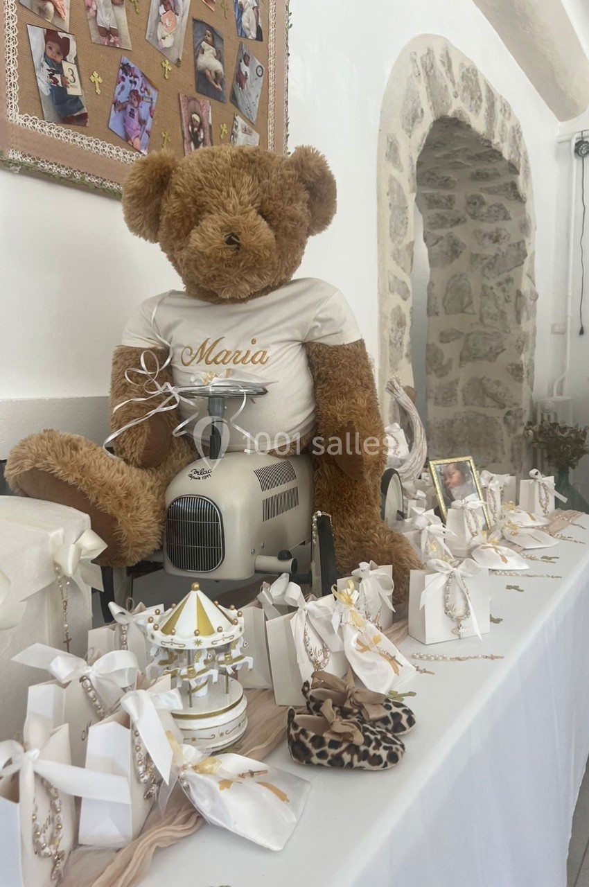 Un ours en peluche sur une machine à coudre, entouré de décorations et de cadeaux sur une table blanche.