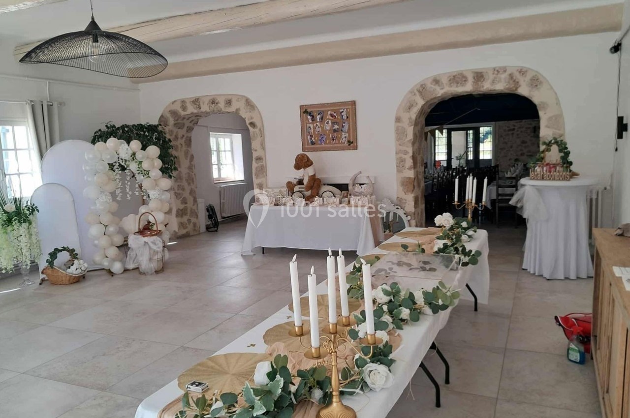 Salle décorée pour un événement avec tables ornées de bougies, fleurs et ballons blancs, dans un cadre en pierre.