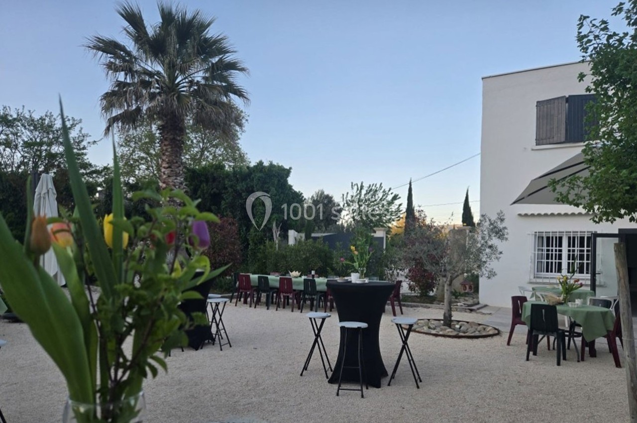 Jardin aménagé avec tables et chaises, palmiers et végétation, devant une maison blanche par temps clair.