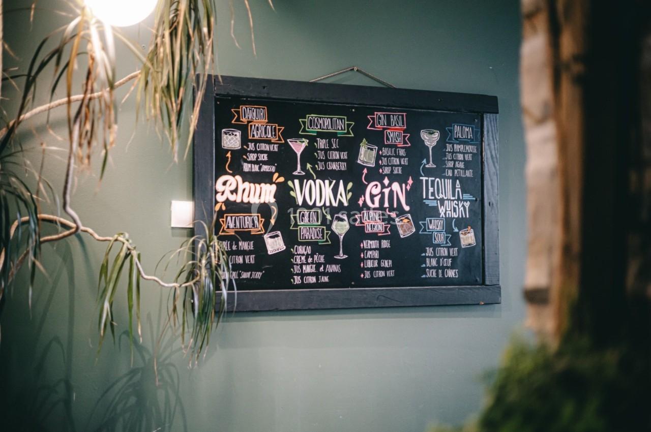 Ardoise murale avec des noms de cocktails et spiritueux écrits à la craie, entourée de plantes.